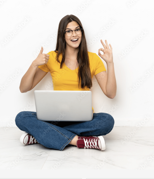 Eine lachende Frau mit gelben T-shirt und Jeans sitzt im Schneidersitz auf dem Boden. Sie hat einen Laptop auf dem Schoß und zeigt mit einer Hand den Daumen nach oben und mit der anderen Hand das 'OK' Zeichen um die Vorteile von Social Media Recruiting zu verdeutlichen.
