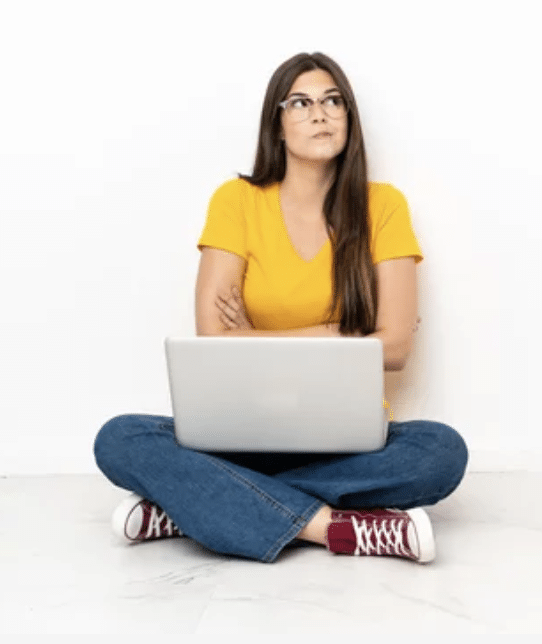 Eine Frau mit gelben T-shirt und Jeans sitzt im Schneidersitz auf dem Boden. Sie hat einen Laptop auf dem Schoß und hat die Arme verschränkt. Dabei schaut sie ernst und fragend nach oben um die Nachteile von Social Media Recruiting zu verdeutlichen.
