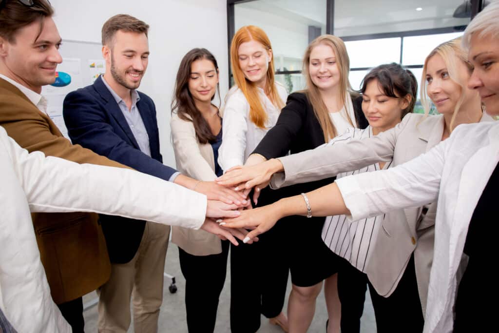 Gruppe eines Business Teams steht freudig im Halbkreis und stapeln jeweils eine Hand übereinander, als Zeichen von Teamwork und Mitarbeiterbindung.