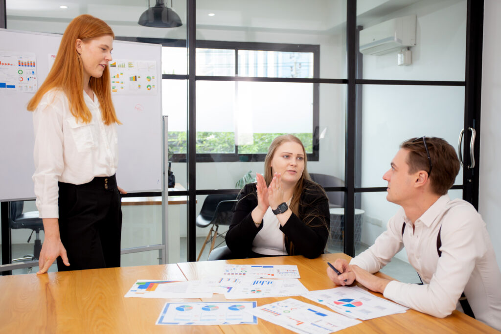 Mitarbeiterbindung messbar machen Zwei Frauen und ein Mann befinden sich an einem Bürotisch. Hinter ihnen ist ein Whiteboard mit Statistiken und auf dem Tisch liegen Dokumente mit Tabellen.