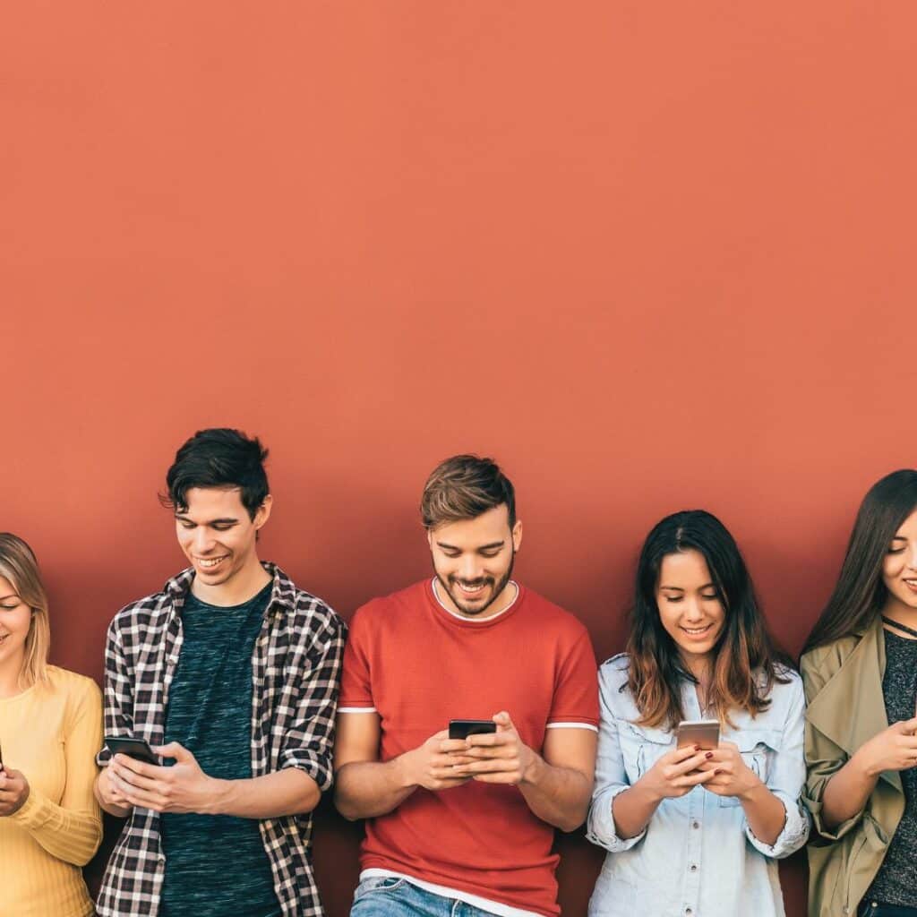 Eine Gruppe junger Menschen vor einer orangenen Wand mit dem Smartphone in der Hand zur Verdeutlichung Moderner Methoden zur Mitarbeitergewinnung