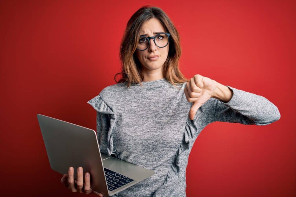 Junge schöne brünette Geschäftsfrau mit Brille, die einen Laptop auf rotem Hintergrund mit wütendem Gesicht benutzt, negatives Zeichen, das Abneigung mit Daumen nach unten zeigt, Ablehnungskonzept, um die Nachteile von Stellenportalen zu zeigen.